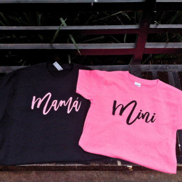 Dúo de playeras 'matching outfit' para madre e hija sobre una banca; camiseta negra con texto 'Mamá' en cursiva rosa y camiseta rosa infantil con texto 'Mini' en cursiva negra.