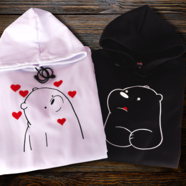 Dúo de sudaderas con capucha para parejas cristianas; una blanca con oso y corazones y una negra con oso serio, estilo matching outfit sobre fondo de madera.