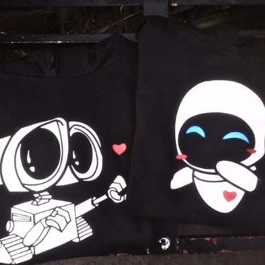 Set de sudaderas negras para parejas cristianas con estampado de WALL-E ofreciendo un corazón y EVE sonriendo; diseño matching outfit sobre banca oscura.